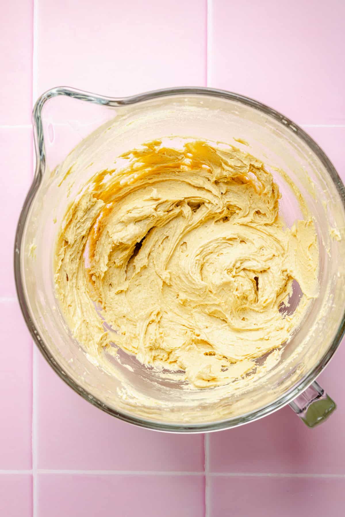 A glass mixing bowl containing creamy, light-yellow cookie dough sits on a pink tiled surface. The dough has been partially mixed, with swirls and streaks visible along the sides of the bowl.