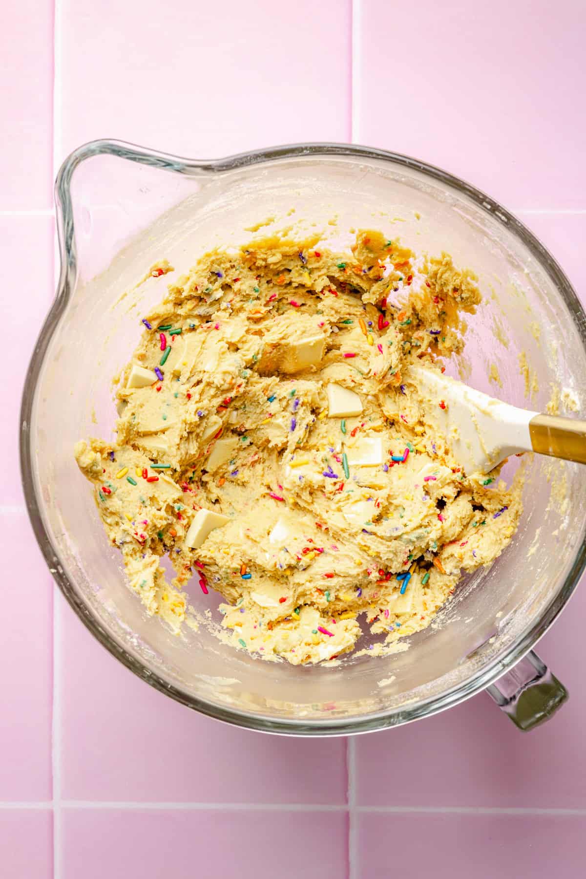 A glass mixing bowl filled with cookie dough, colorful sprinkles, and chunks of butter sits on a pink tiled surface. A spatula with a gold handle rests in the dough.