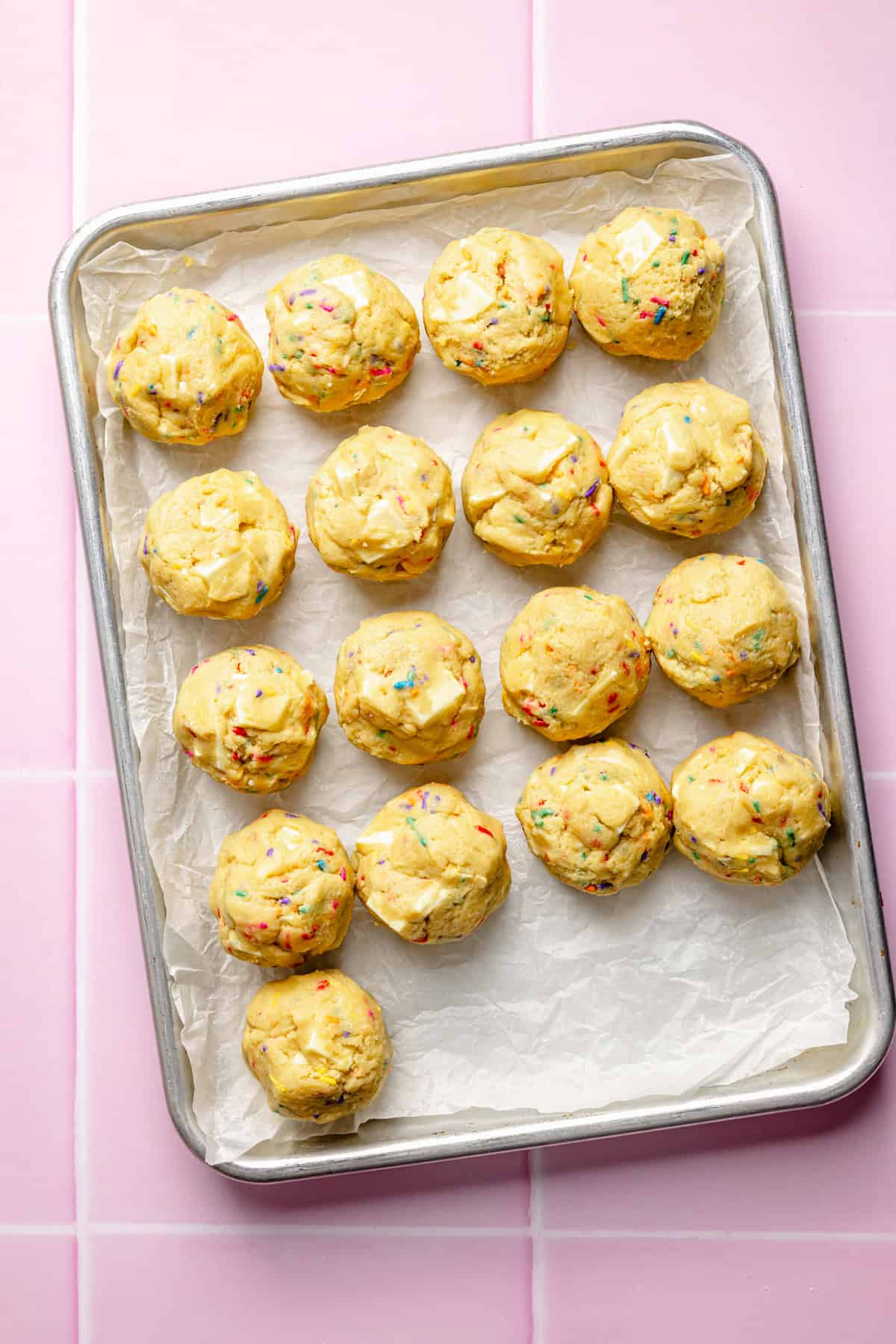 A baking tray lined with parchment paper holds sixteen round, unbaked funfetti cookie dough balls, placed neatly in rows, on a pink tiled surface.