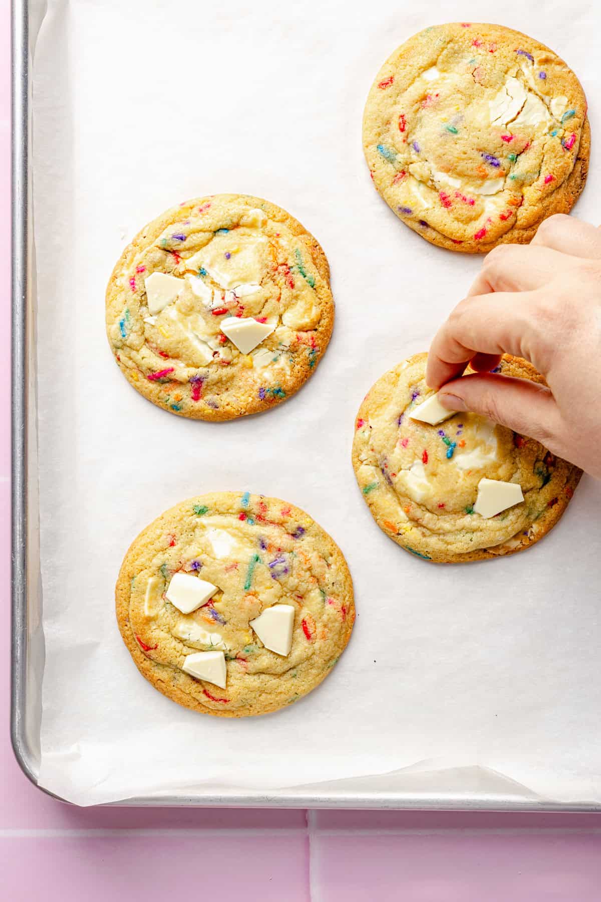 A hand adds white chocolate pieces on top of large, round cookies with colorful sprinkles, arranged on a parchment-lined baking sheet.