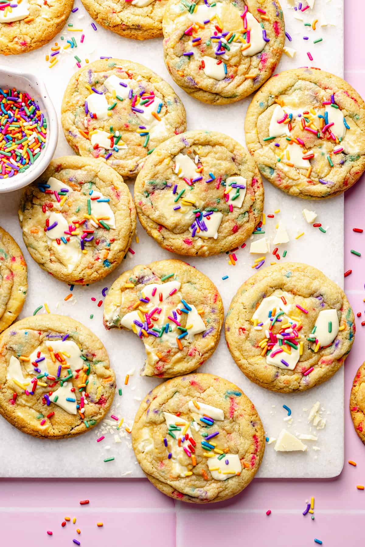 A batch of colorful birthday cake cookies topped with white chocolate chunks and rainbow sprinkles is arranged on a white surface. One cookie has a bite taken out of it, and a small bowl of sprinkles sits nearby.