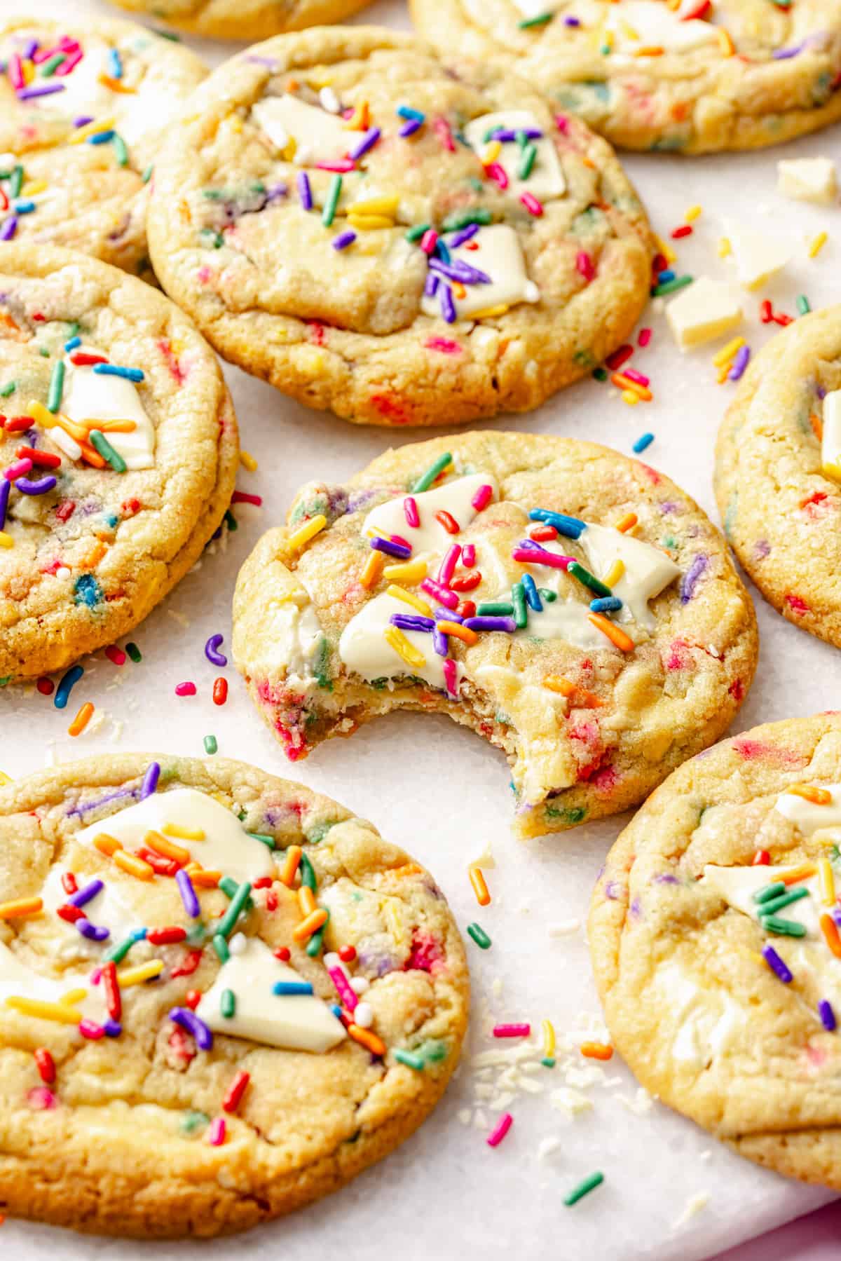 Cookies topped with white chocolate pieces and colorful sprinkles are arranged on parchment paper. One cookie has a bite taken out of it, showing a soft interior. Sprinkles are scattered around the cookies.