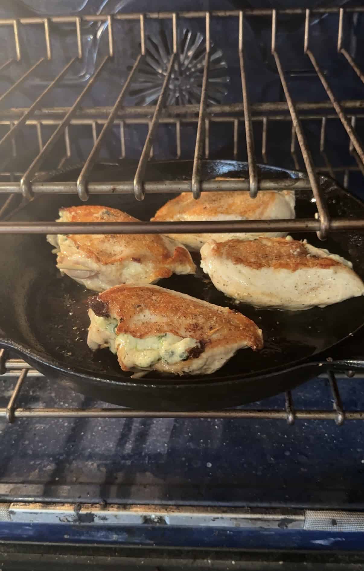 Four pieces of boursin stuffed chicken breast with golden-brown skin are cooking in a black cast iron skillet inside an oven. The chicken appears seasoned and is being baked.