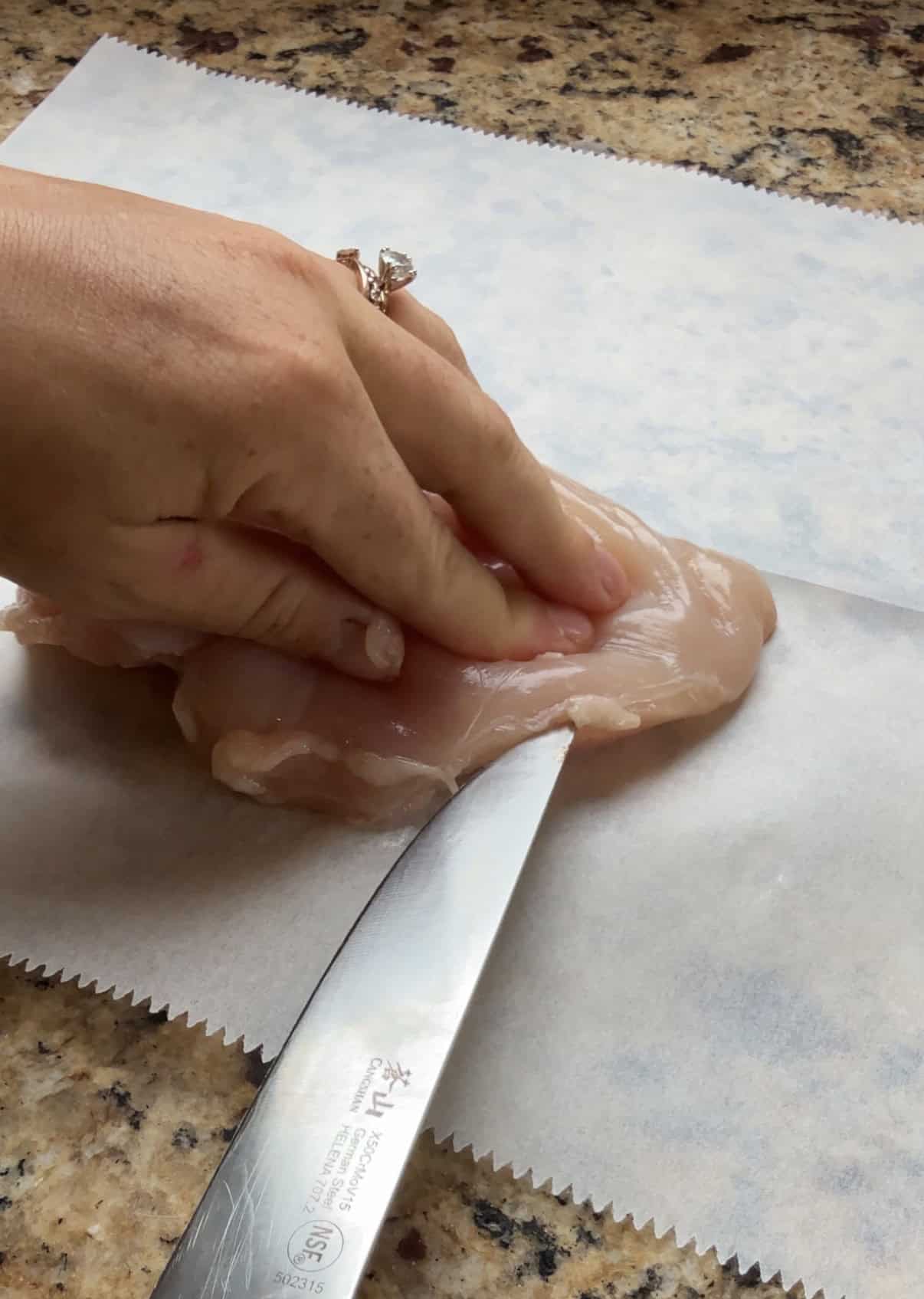 A hand holds a raw chicken breast on parchment paper while slicing it horizontally with a knife on a kitchen counter.