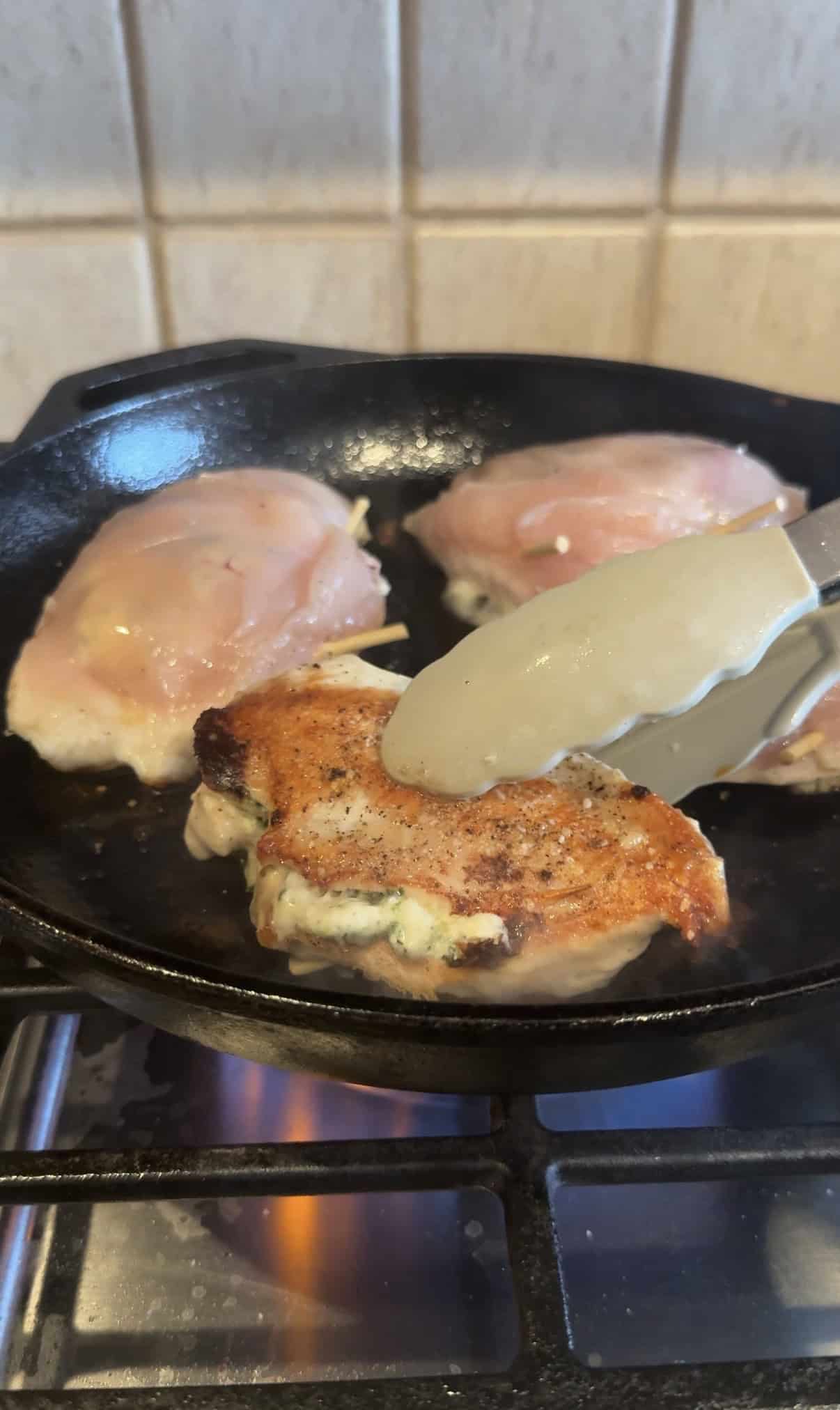 Three stuffed chicken breasts cooking in a black cast iron skillet on a stove. One chicken breast is being flipped with tongs, showing its browned, crispy side. The background shows a tiled kitchen wall.