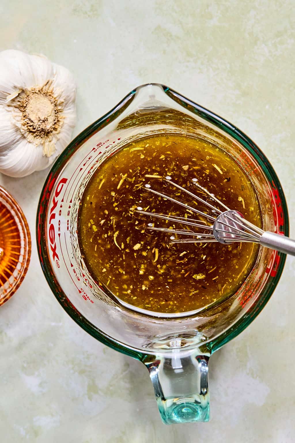 A glass measuring cup filled with a balsamic marinade for balsamic chicken salad is being whisked, with visible garlic and herbs. Nearby, a whole garlic bulb and a small bowl of brown liquid rest on a light surface.