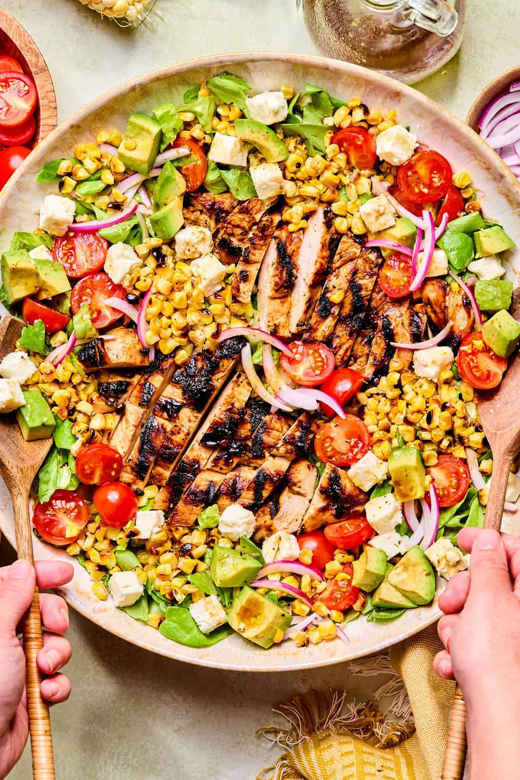 A large bowl of balsamic chicken salad with avocado, cherry tomatoes, corn, red onions, lettuce, and feta cheese is served with wooden salad tongs.