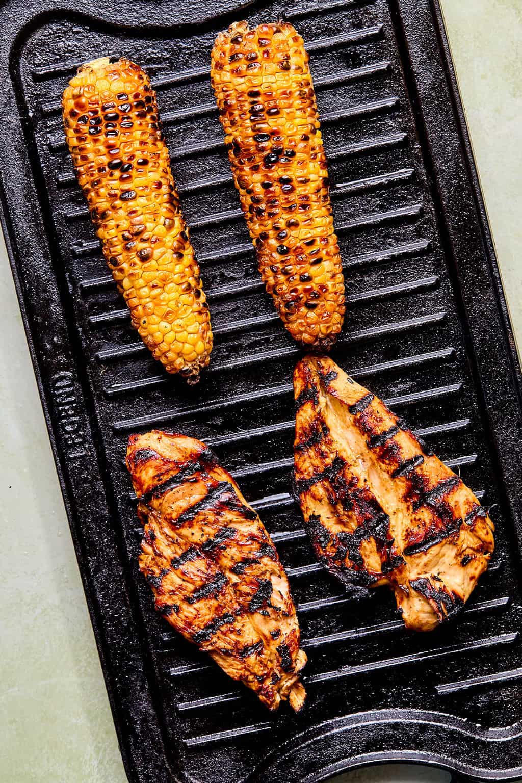 Two grilled corn cobs and two grilled chicken breasts with visible char marks on a black grill pan—perfect for a hearty balsamic chicken salad.