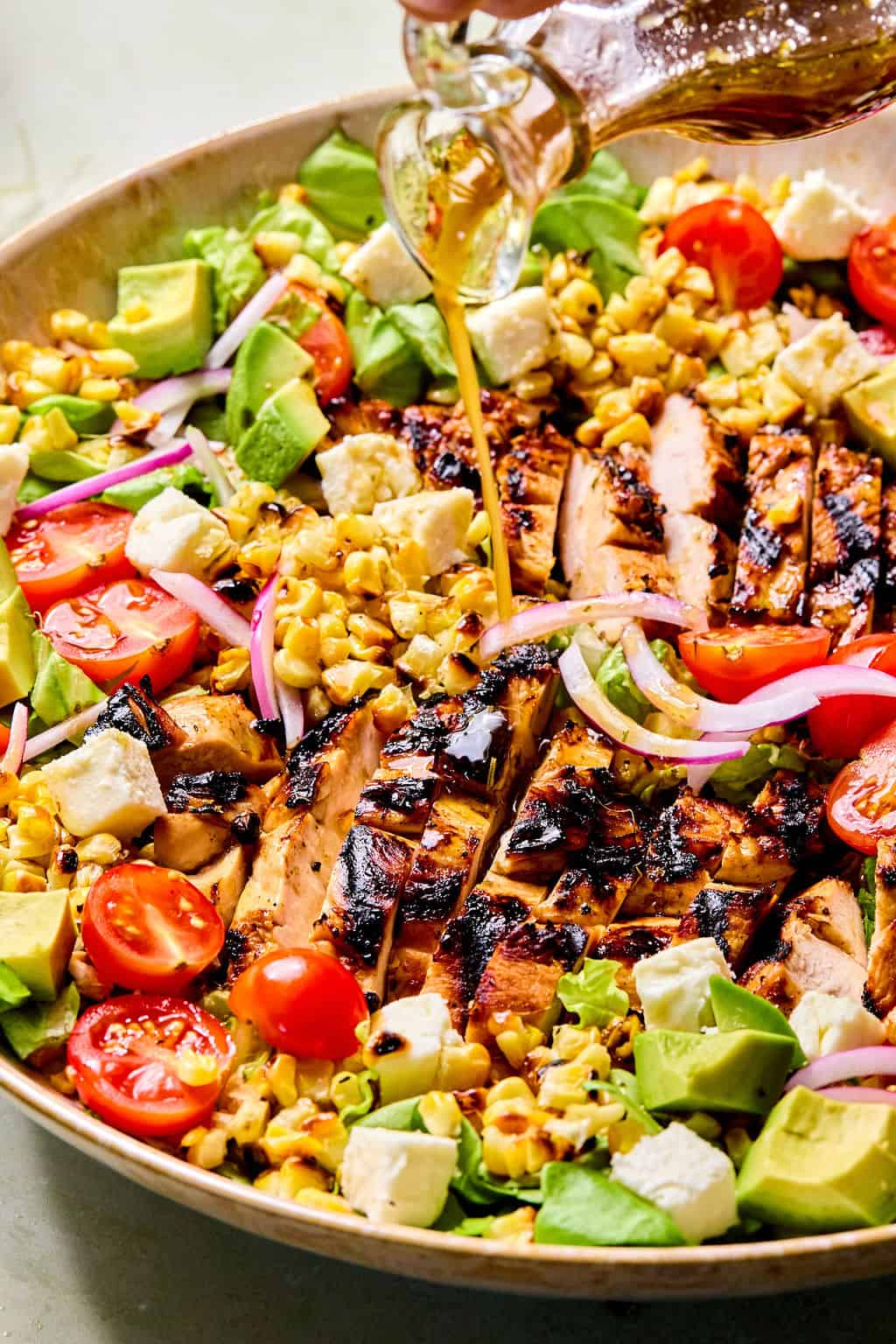 A close-up of a salad bowl with balsamic marinated grilled chicken slices, cherry tomatoes, avocado, corn, red onion, and feta cheese, while a hand pours balsamic dressing over the top.