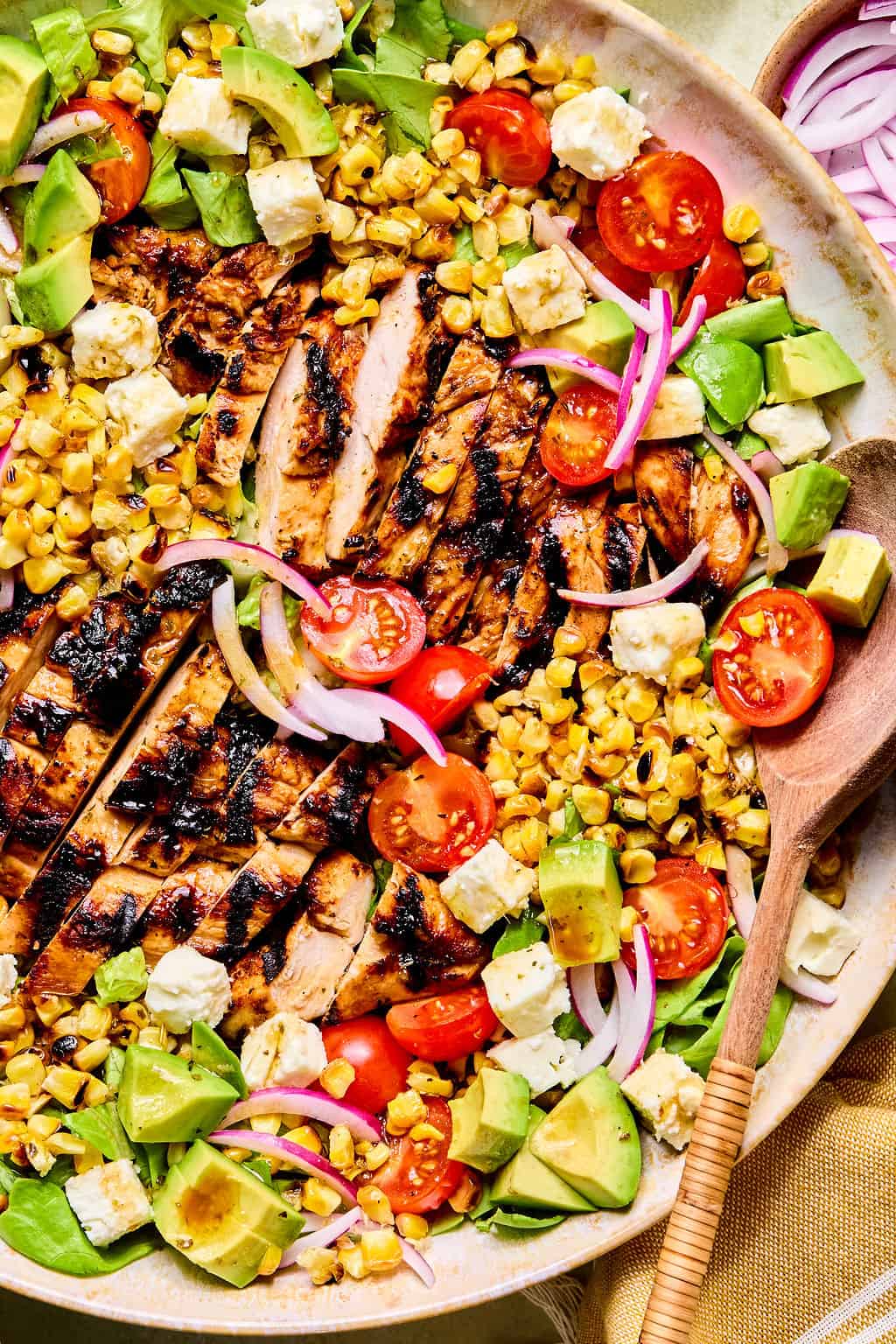 A colorful balsamic chicken salad with grilled sliced chicken, avocado, cherry tomatoes, grilled corn, red onion, feta cheese, and greens, served in a bowl with a wooden spoon.