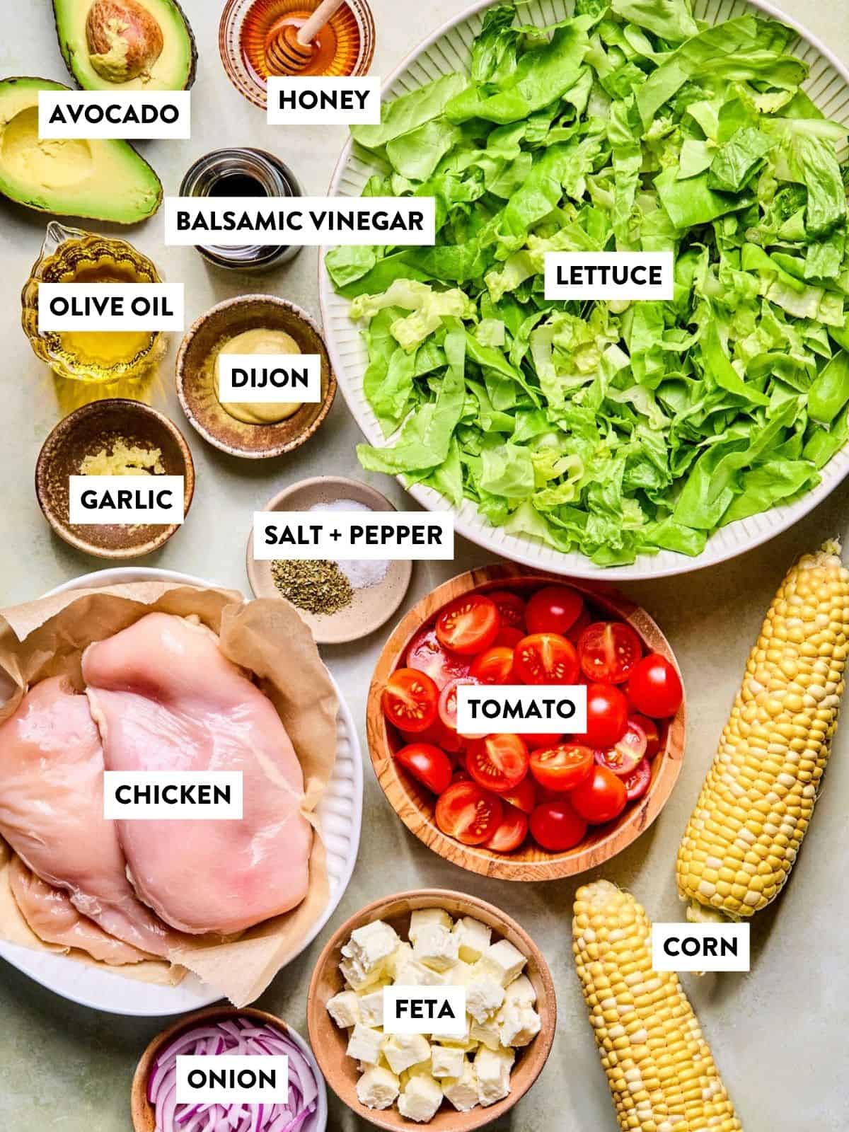 Top-down view of salad ingredients: chopped lettuce, cherry tomatoes, corn cobs, chicken breasts, avocado halves, feta cheese, sliced red onion, garlic cloves, salt and pepper, Dijon mustard, olive oil, balsamic vinegar, and honey.