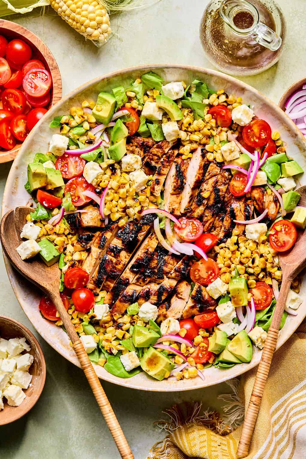 A large bowl of balsamic chicken salad with corn, avocado, cherry tomatoes, sliced red onions, and feta cheese is served with wooden utensils. Nearby are a bowl of tomatoes, feta, corn, and a glass jug of dressing.