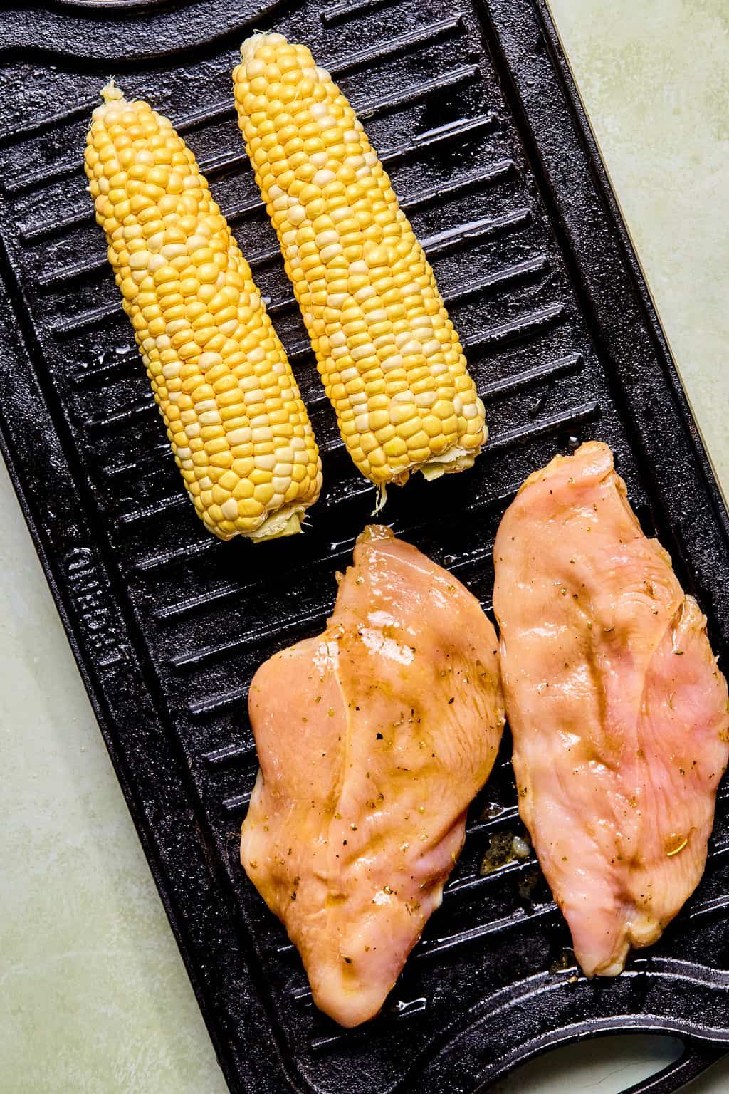 Two raw chicken breasts and two ears of raw corn on the cob are placed side by side on a black grill pan, ready to be cooked for a fresh balsamic chicken salad.