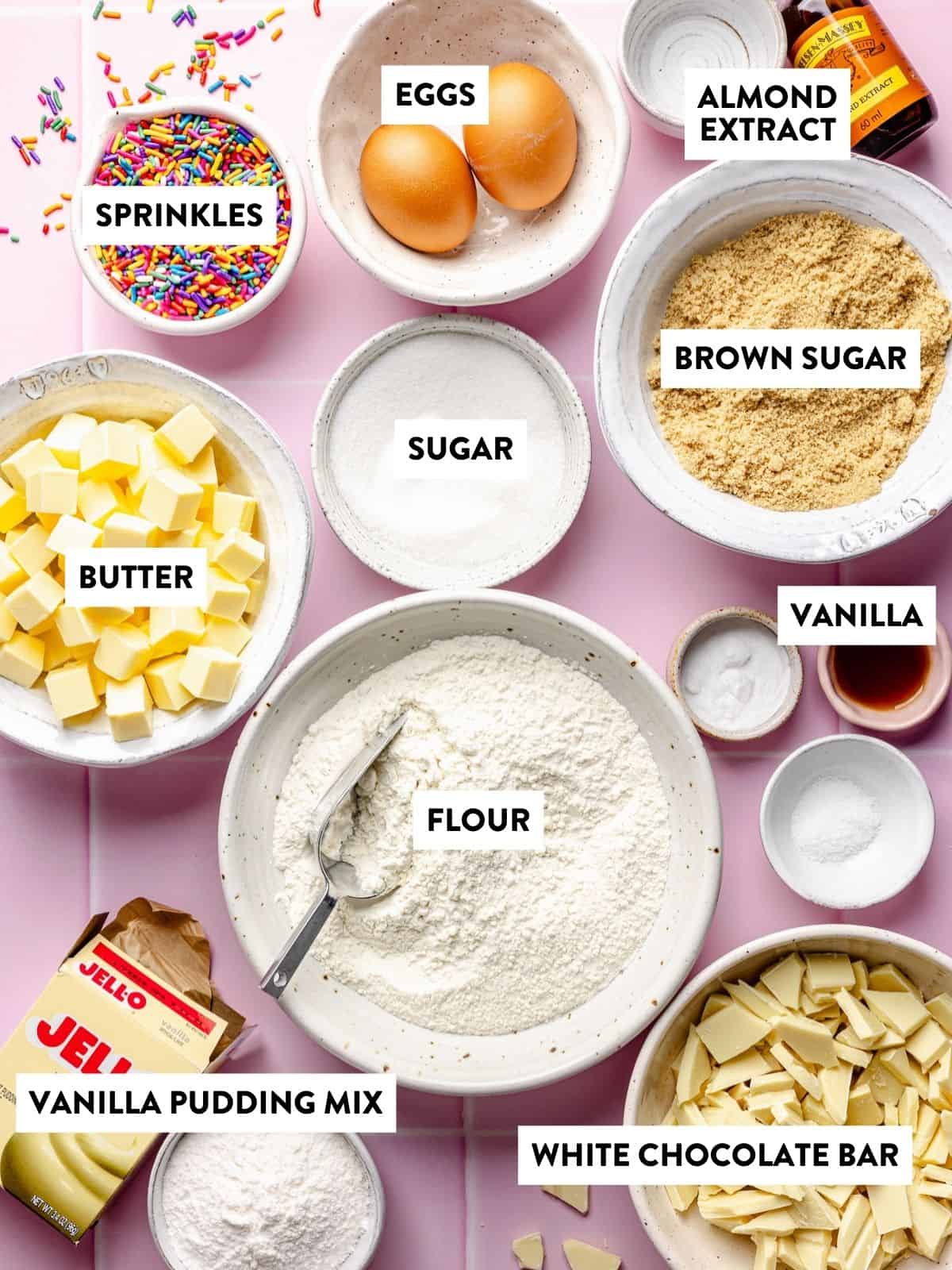 A flat lay of baking ingredients in bowls on a pink surface, including labeled butter, eggs, flour, sugar, brown sugar, almond extract, vanilla, sprinkles, white chocolate bar, and vanilla pudding mix.