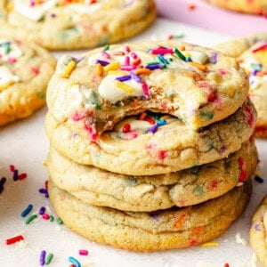A stack of four birthday cake cookies with colorful sprinkles and white chocolate chips. The top cookie has a bite taken out, and more cookies and sprinkles are scattered in the background.