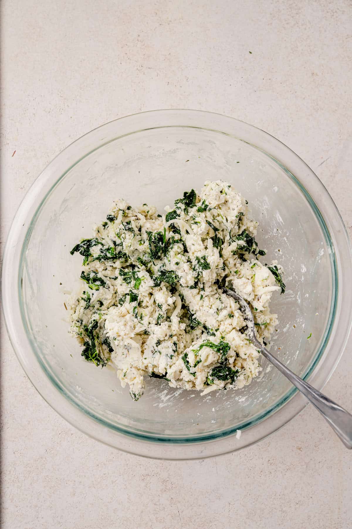 A glass bowl filled with a mixture of boursin cheese, spinach, and herbs, with a metal spoon resting inside, on a light-colored surface.