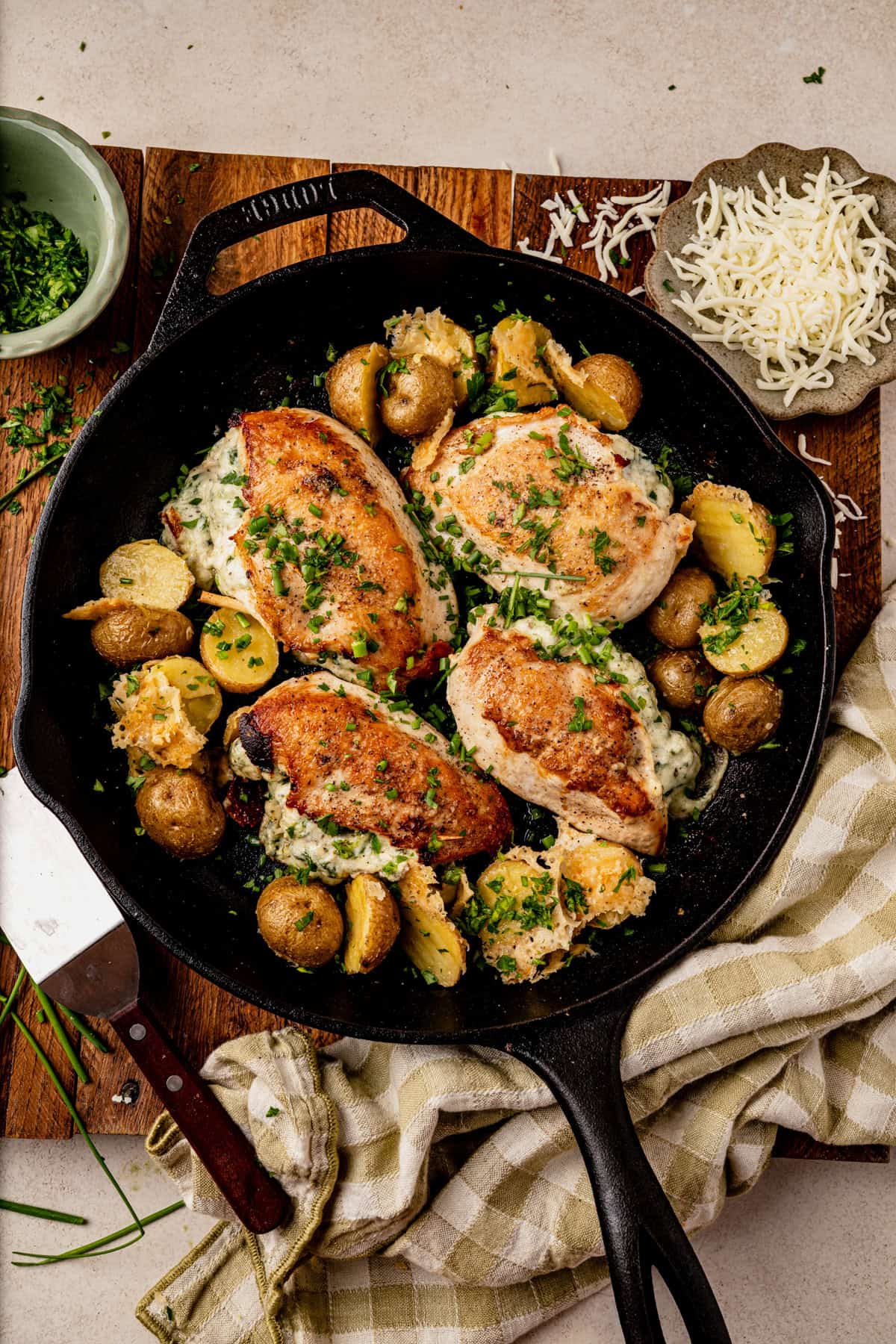 A cast-iron skillet with four golden-brown Boursin stuffed chicken breasts, melted cheese, baby potatoes, and chopped herbs, resting on a wooden board with a cheese grater and a bowl of shredded cheese nearby.