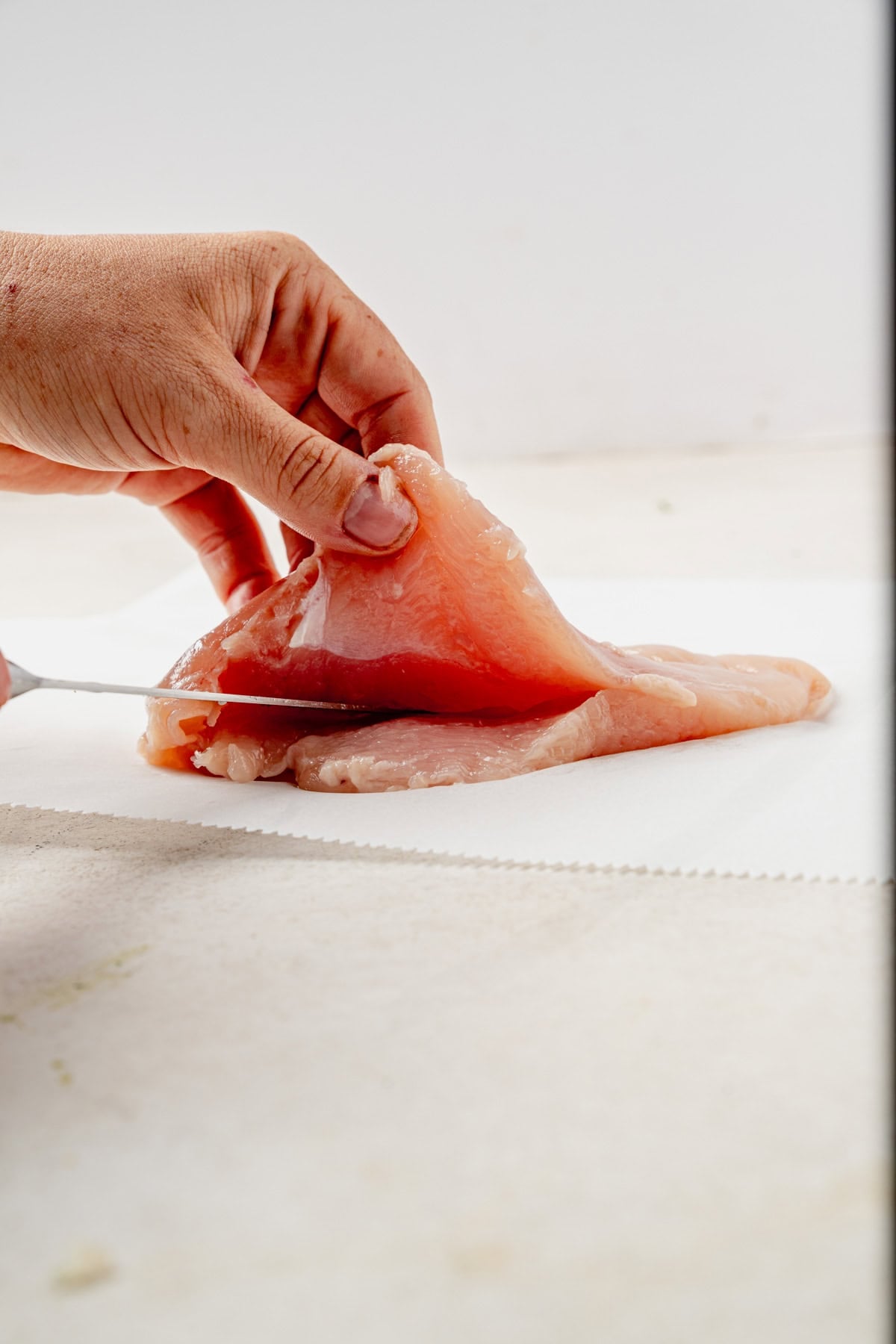 A hand holds a raw chicken breast while slicing it with a knife on a white surface.