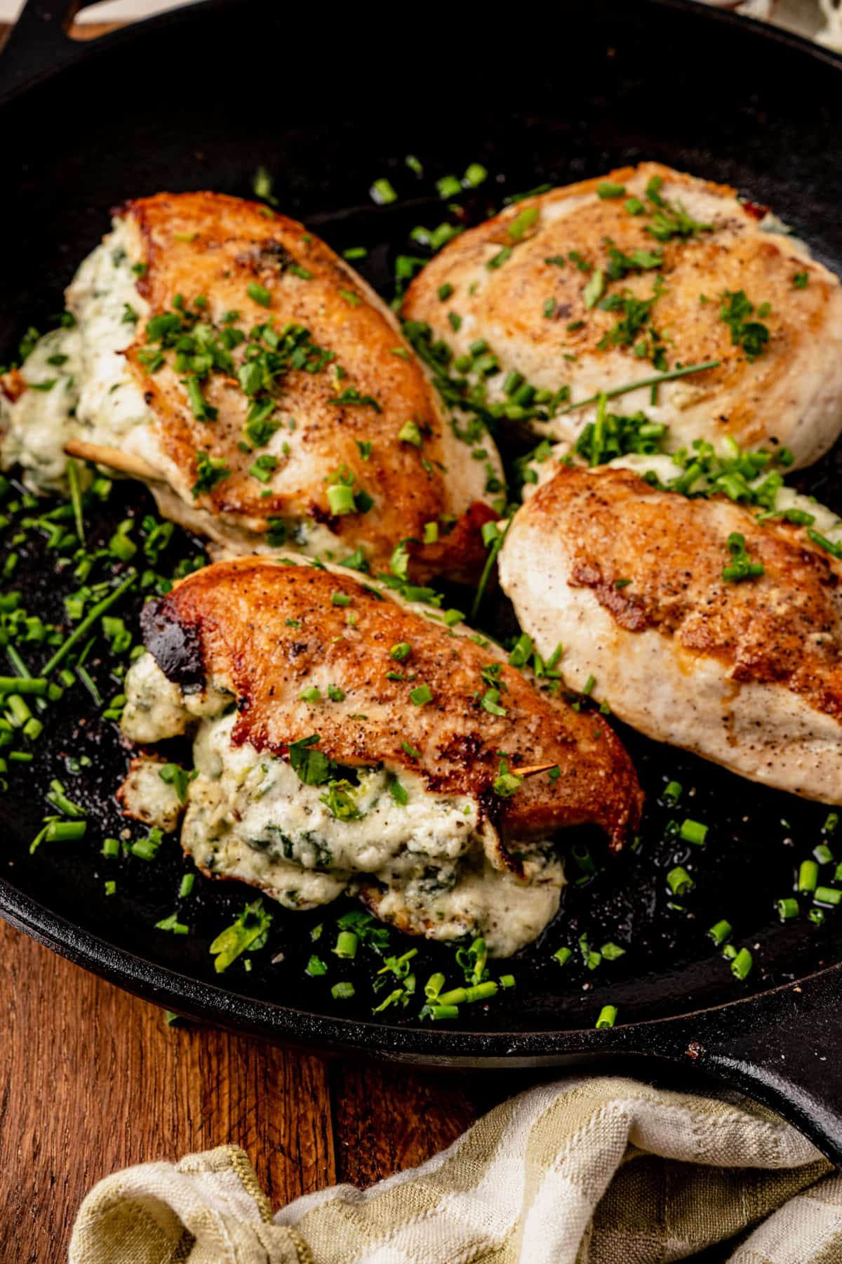 Four golden-brown, pan-seared boursin stuffed chicken breasts with creamy cheese and herbs, garnished with chopped chives, are shown in a black skillet on a wooden surface.