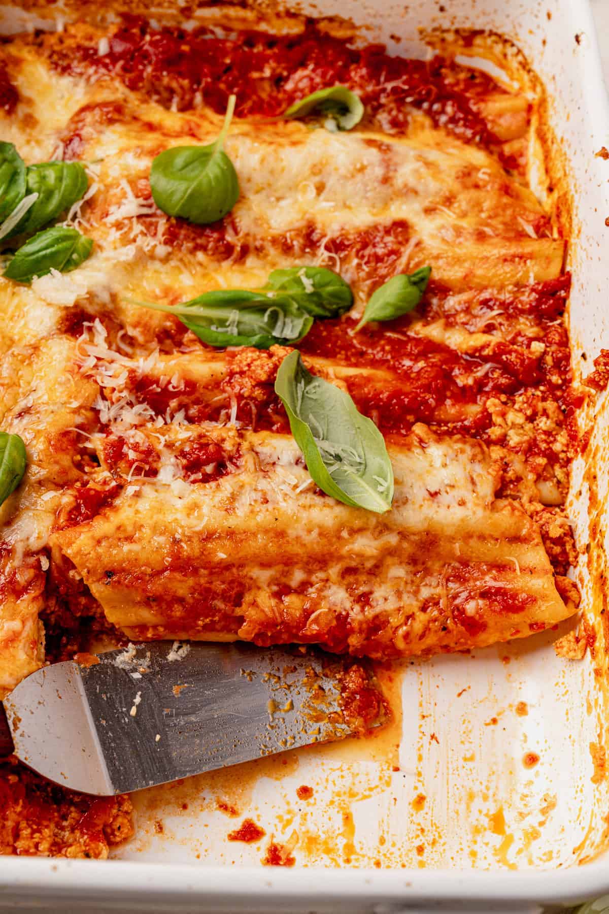 Baked manicotti filled with cheese, topped with tomato sauce, melted cheese, and fresh basil leaves in a white baking dish. A serving spatula rests in the dish.