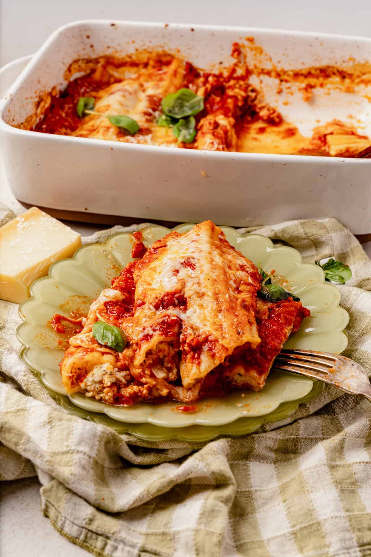 A serving of cheesy, baked stuffed manicotti topped with tomato sauce and basil sits on a green plate, with a fork beside it. In the background, a baking dish of more pasta and a wedge of cheese are visible.