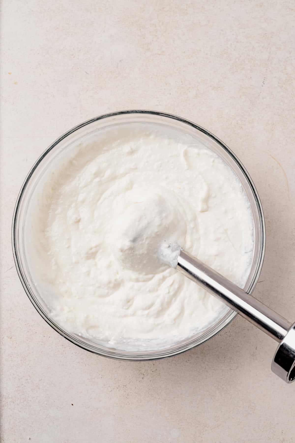 A glass bowl filled with blended cottage cheese with an immersion blender partially submerged in the mixture on a light-colored surface.