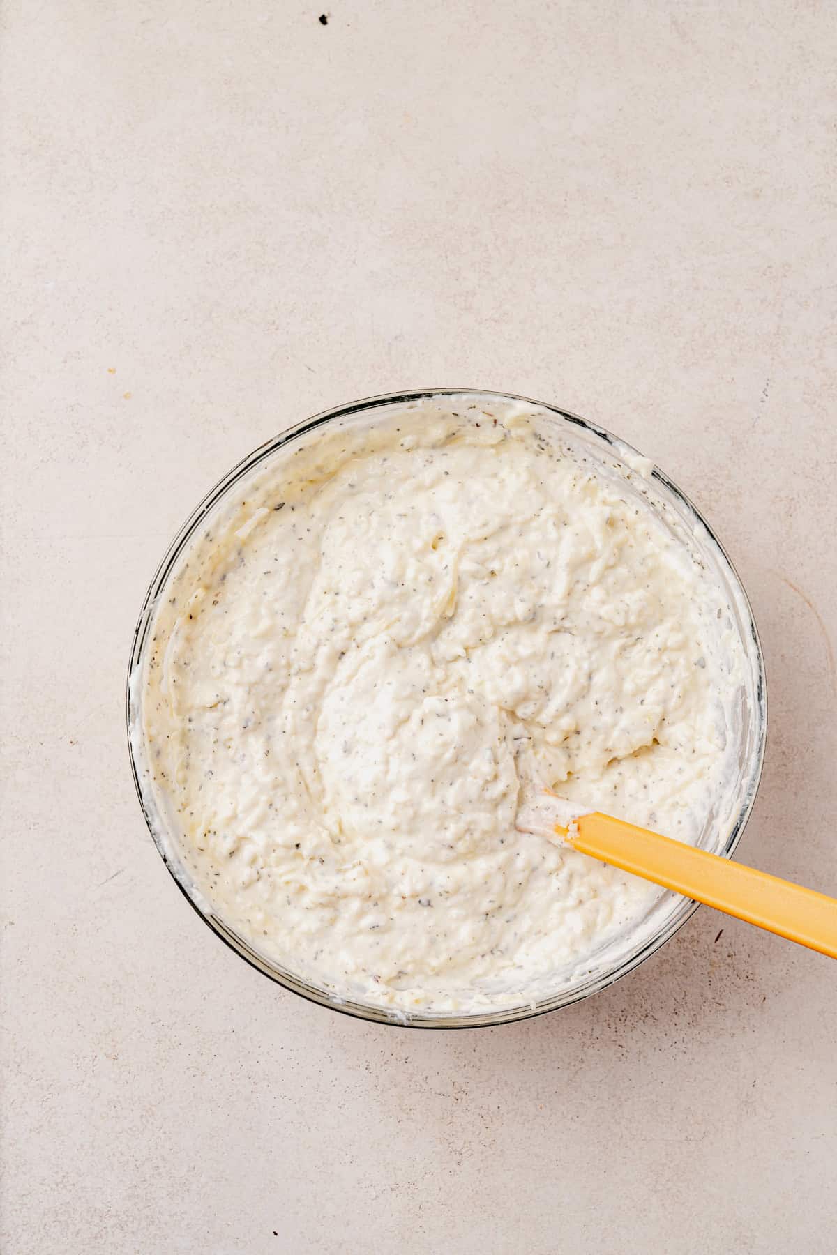 A glass bowl filled with a creamy, white blended cottage cheese mixture speckled with herbs, sitting on a light-colored surface. A yellow spatula rests in the bowl, partially submerged in the mixture.