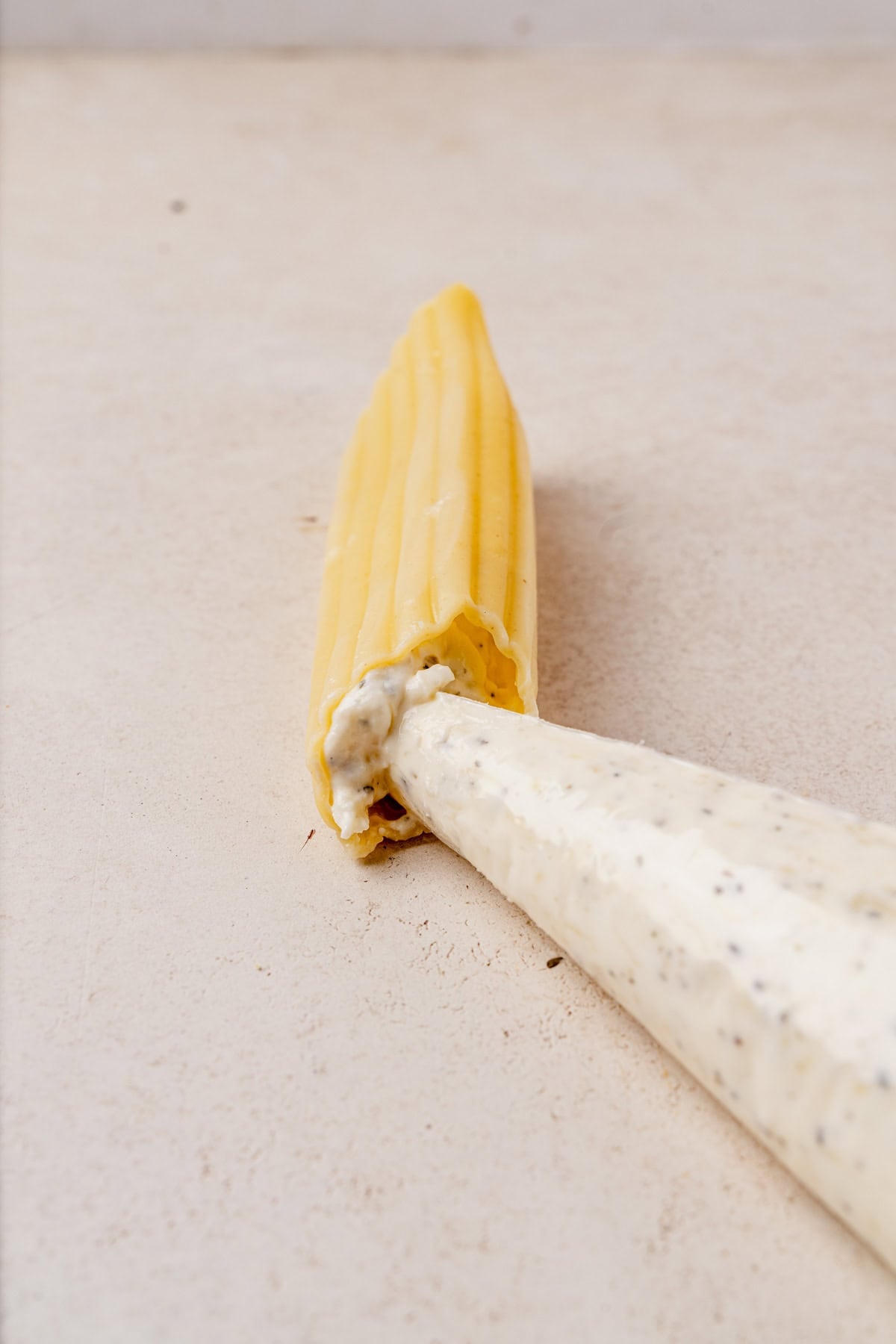 A piping bag filled with creamy cottage cheese mixture is being used to stuff a large cooked manicotti pasta shell on a light-colored surface.
