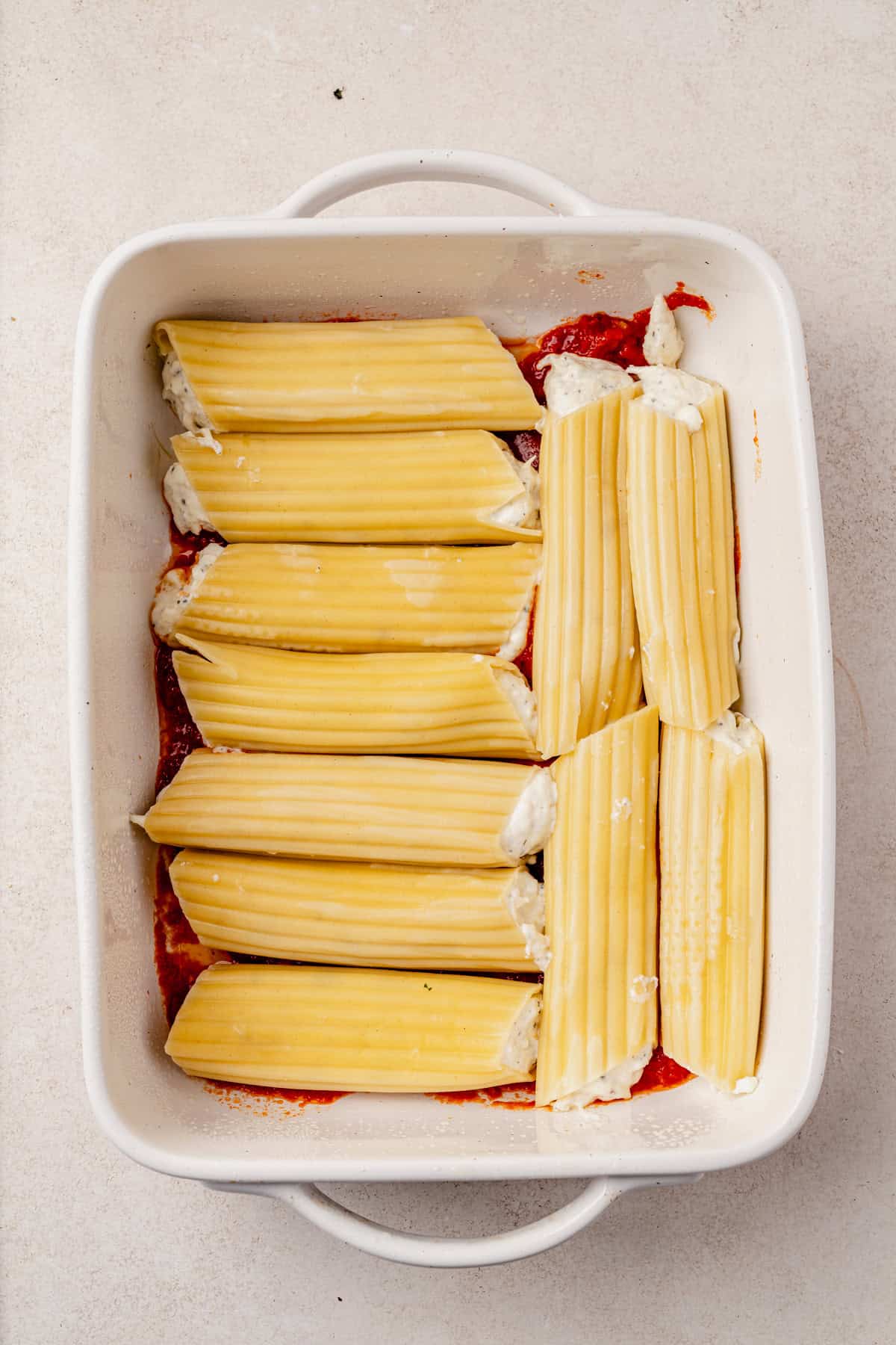 A white baking dish filled with uncooked manicotti pasta stuffed with ricotta cheese, placed over a layer of tomato sauce on a light-colored surface.