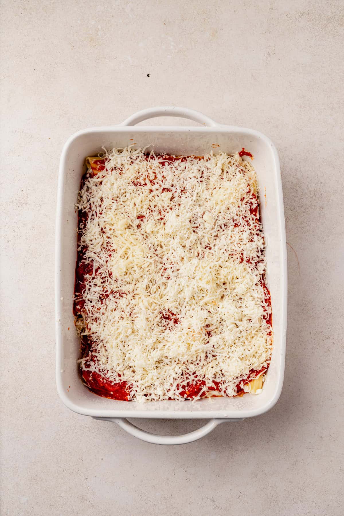 A white rectangular baking dish filled with uncooked manicotti, topped with tomato sauce and a generous layer of shredded cheese, sits on a light-colored surface.
