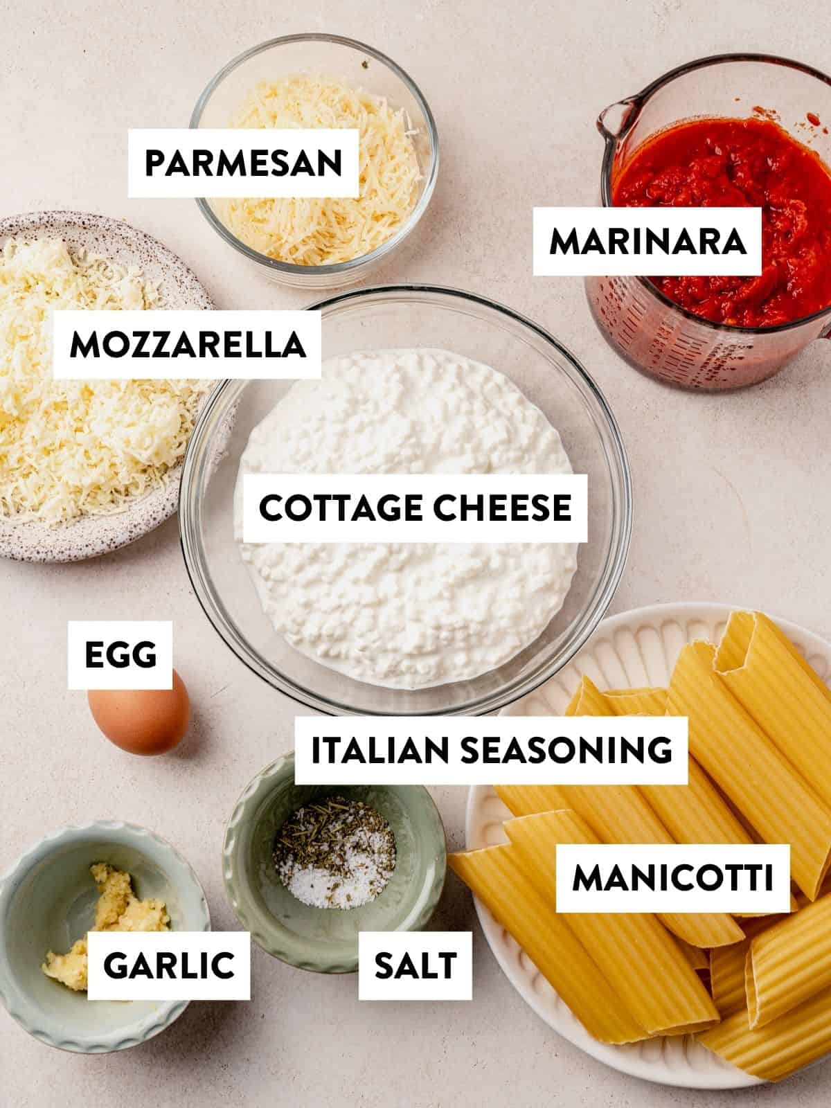 Bowls and containers hold labeled ingredients for manicotti: parmesan, mozzarella, cottage cheese, marinara sauce, an egg, Italian seasoning, minced garlic, salt, and uncooked manicotti pasta.