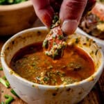 A hand dips a piece of steak coated in cowboy butter into a bowl of melted butter, with green onions scattered nearby.