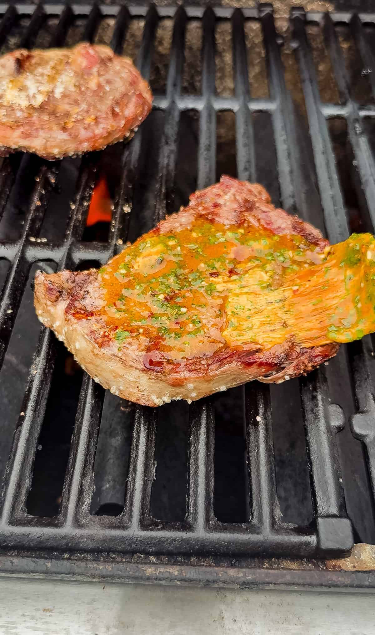 A piece of steak topped with cowboy butter is cooking on a grill, with visible grill marks and small flames beneath the grates. Another piece of meat is partially visible in the background.