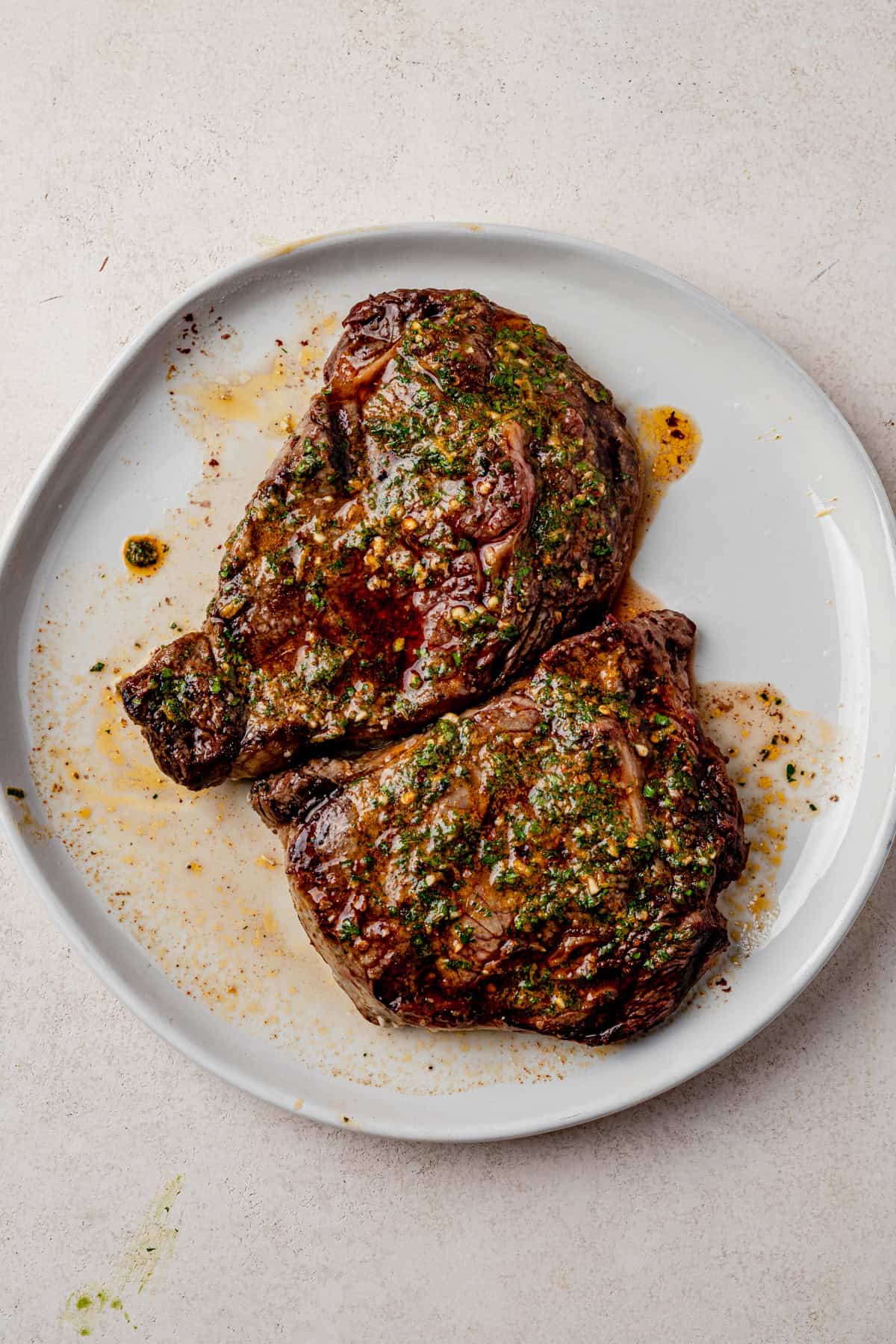 Two cooked steaks topped with cowboy butter are served on a white plate. Juices and seasoning are visible around the steaks against a light-colored background.