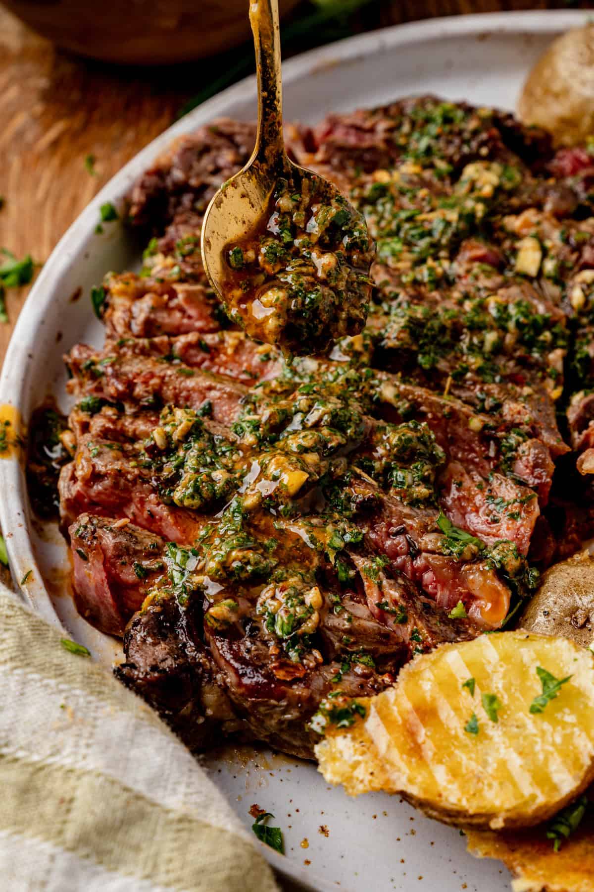 A close-up of a juicy grilled steak topped with cowboy butter, with a spoon drizzling more sauce on top and slices of seasoned potato on the side.