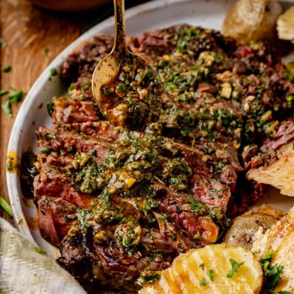 Sliced steak on a plate topped with melted cowboy butter and a hint of cowboy butter, as a spoon drizzles more sauce over it. Roasted potatoes and herbs are visible on the side.