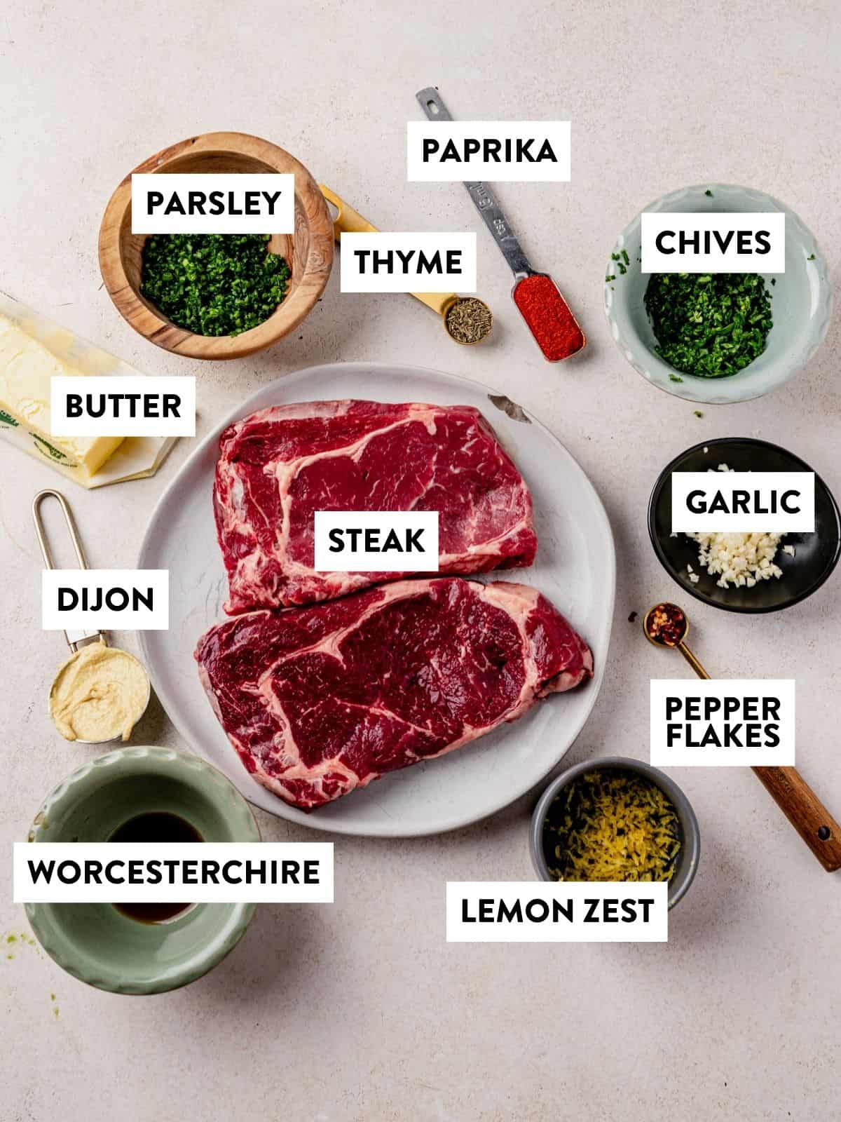 Two raw steaks on a plate surrounded by labeled ingredients: chopped parsley, thyme, paprika, chives, minced garlic, red pepper flakes, lemon zest, Worcestershire sauce, Dijon mustard, and butter.