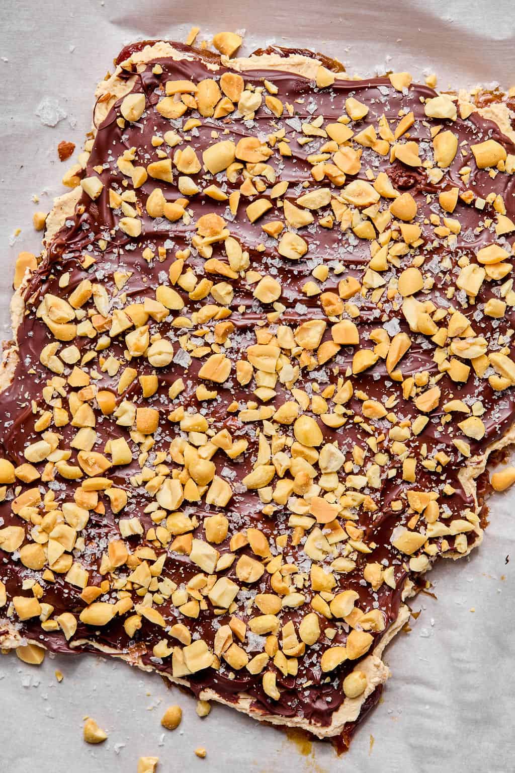 A slab of date bark topped with chopped peanuts and a sprinkle of flaky sea salt, spread on parchment paper.