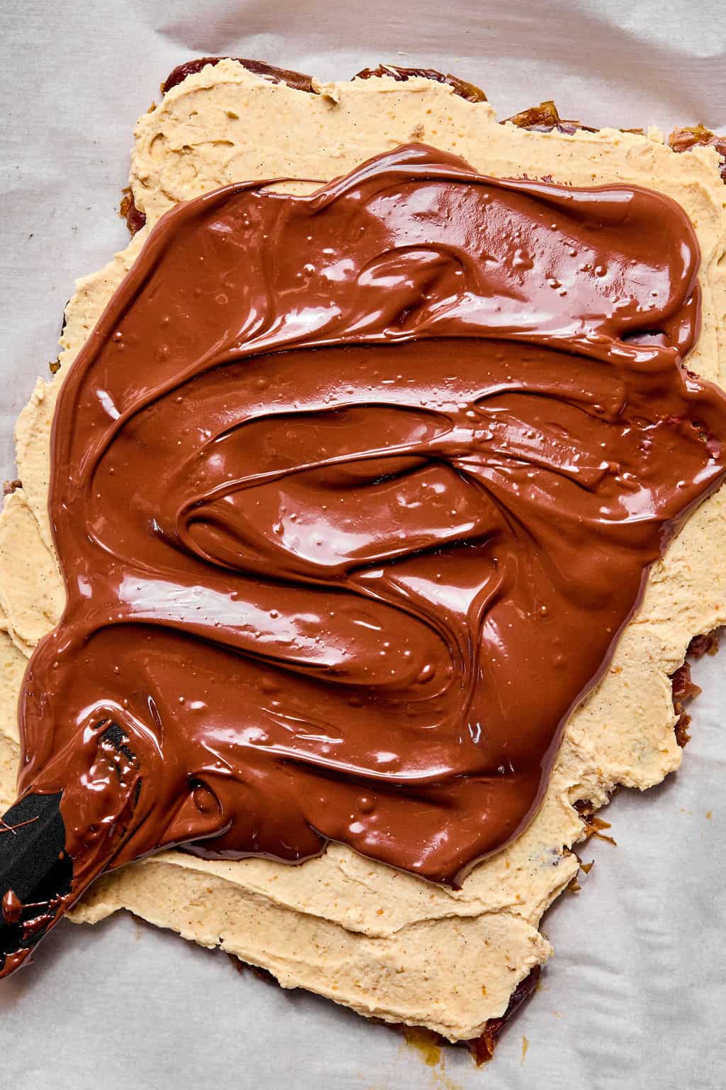 A spatula spreads a thick layer of glossy melted chocolate over a rectangular base topped with a peanut butter yogurt layer, all set on parchment paper.