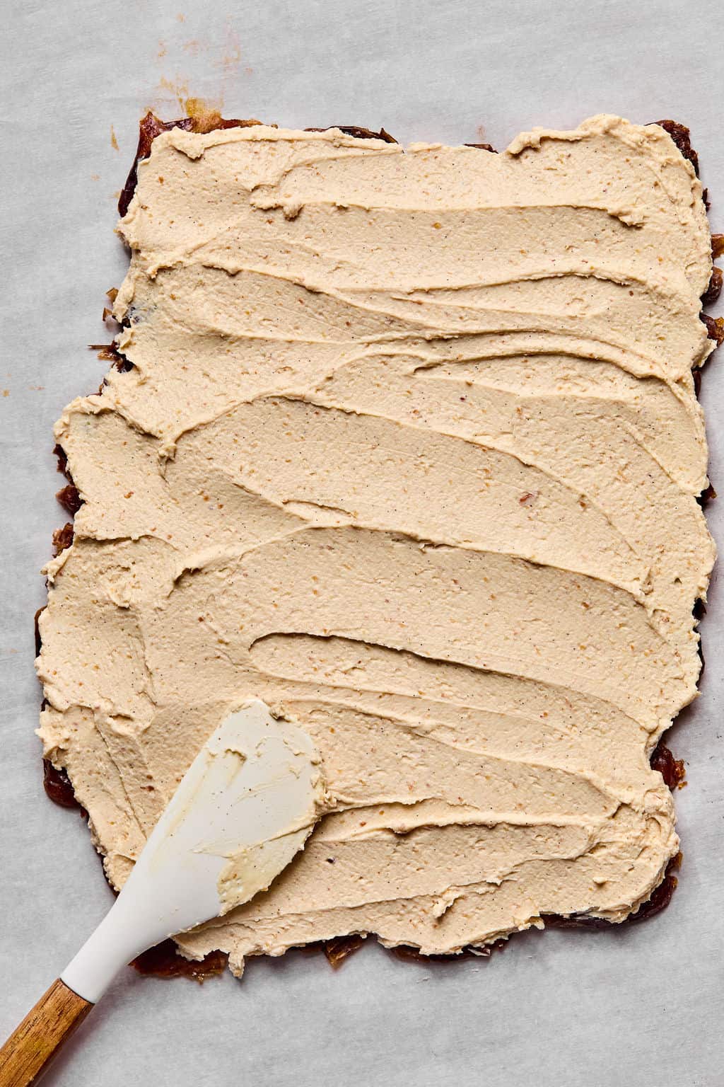 A rectangular sheet of dates on parchment paper is being covered with a thick, even layer of greek yogurt and peanut butter mixture spread with a white spatula with a wooden handle.
