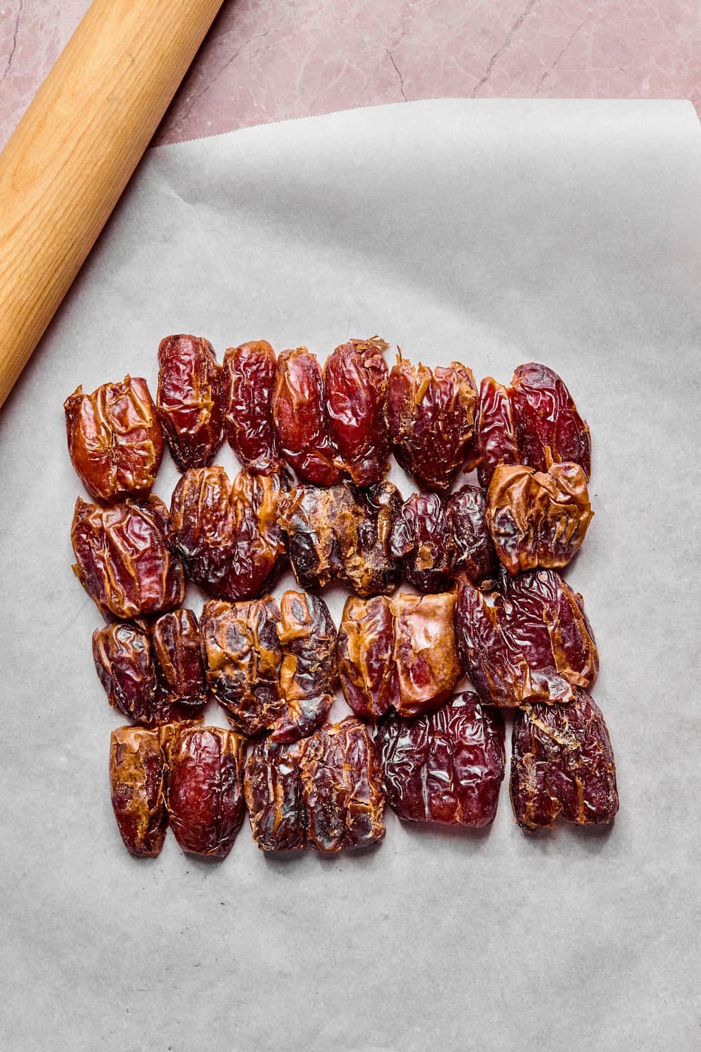 Twenty whole dates are arranged in neat rows on a sheet of parchment paper, next to a wooden rolling pin, all set on a pink marble surface.