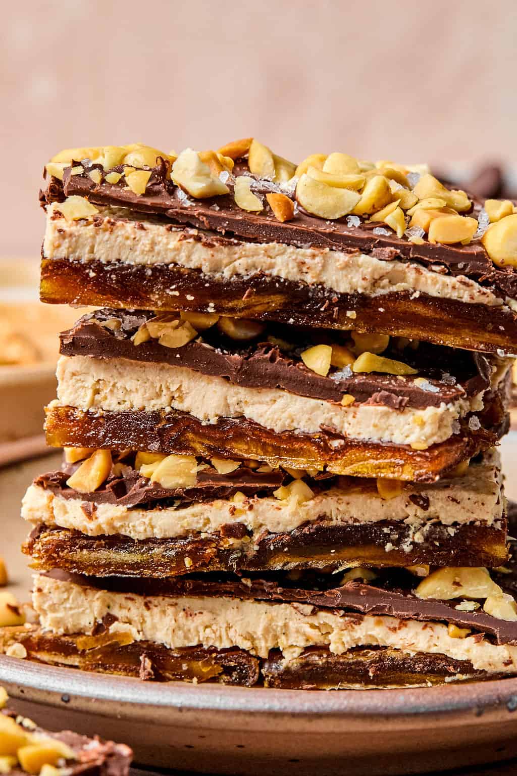 A close-up of four stacked homemade date bark recipe featuring layers of dates, peanut butter, chocolate, and chopped peanuts, served on a ceramic plate with scattered peanut pieces around.