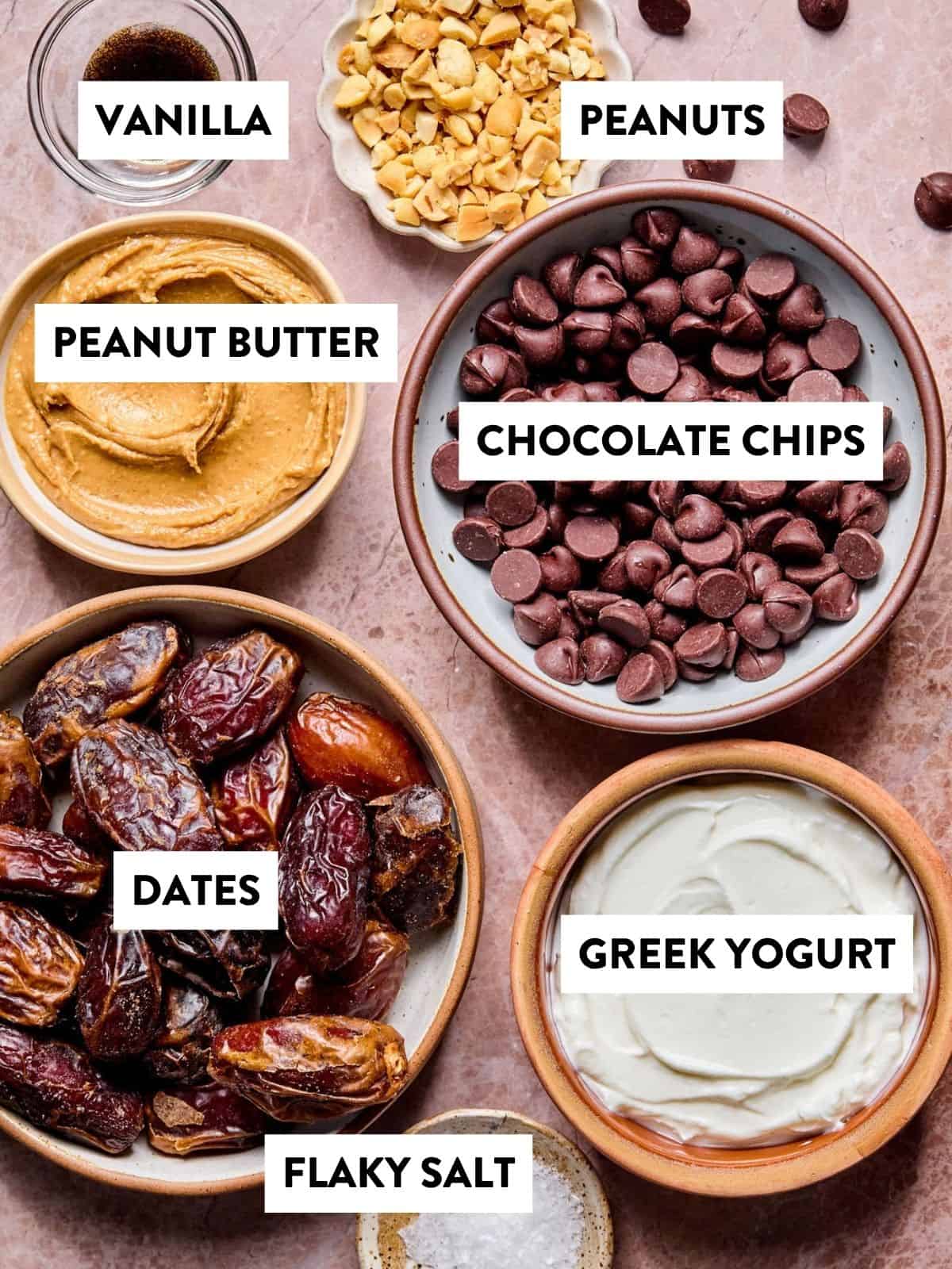 Bowls containing vanilla, chopped peanuts, peanut butter, chocolate chips, dates, Greek yogurt, and flaky salt are arranged on a pink marble surface, each labeled with bold text.