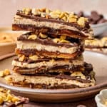 A stack of date bark topped with chopped peanuts and chocolate sits on a ceramic plate. The bars have visible layers of dates, peanut butter, and chocolate. Some peanuts and chocolate pieces are scattered nearby.