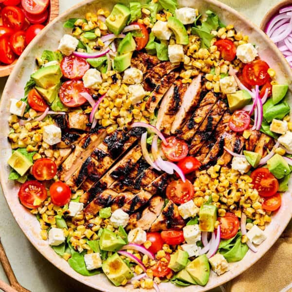 A colorful balsamic chicken salad with grilled chicken slices, cherry tomatoes, avocado, corn, red onion, feta cheese, and lettuce, arranged on a large round plate. Small bowls of cherry tomatoes and red onion are beside the plate.