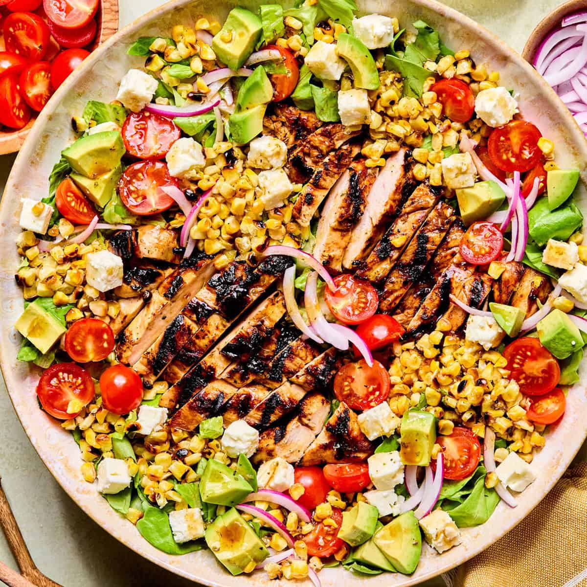 A colorful balsamic chicken salad with grilled chicken slices, cherry tomatoes, avocado, corn, red onion, feta cheese, and lettuce, arranged on a large round plate. Small bowls of cherry tomatoes and red onion are beside the plate.