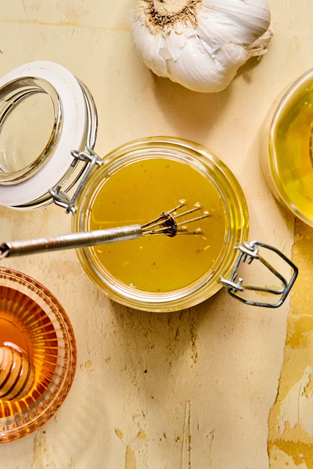 A jar of honey lime vinaigrette with a small whisk inside sits on a light surface, surrounded by a head of garlic, a bowl of honey, and oil—perfect for drizzling over your fresh mango avocado salad.