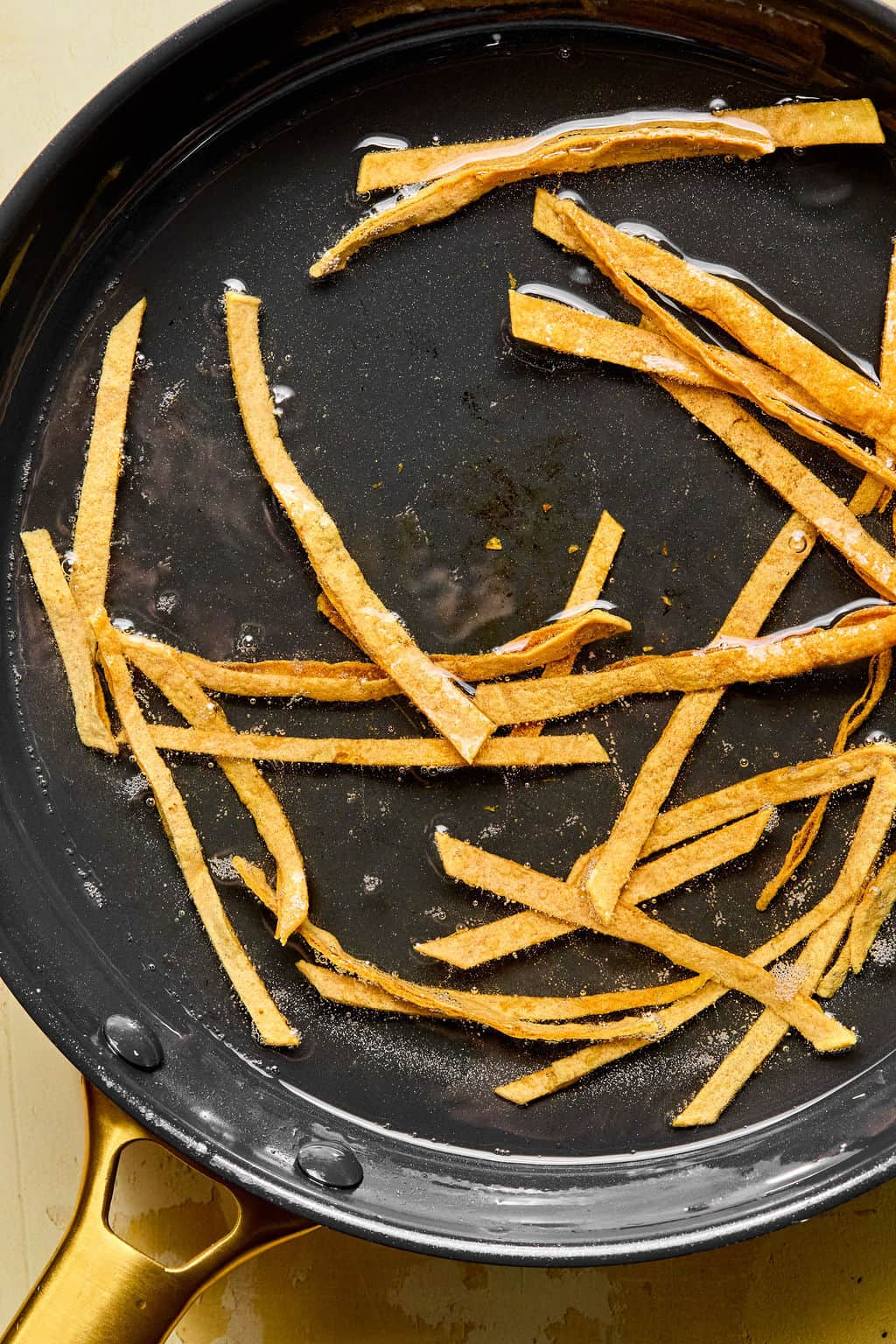 Thin strips of tortilla are being fried in oil in a black skillet with a gold handle. The golden brown, crisping tortilla strips are perfect for topping a fresh mango avocado salad.