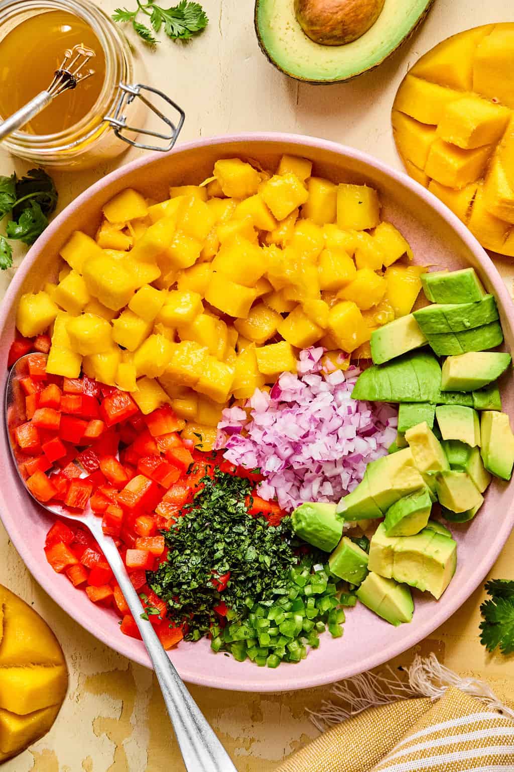 A vibrant mango avocado salad is showcased in a bowl, with diced mango, avocado, red onion, red bell pepper, cilantro, and jalapeño arranged in sections. A spoon rests inside, surrounded by fresh mango, avocado, and a jar of vinaigrette.