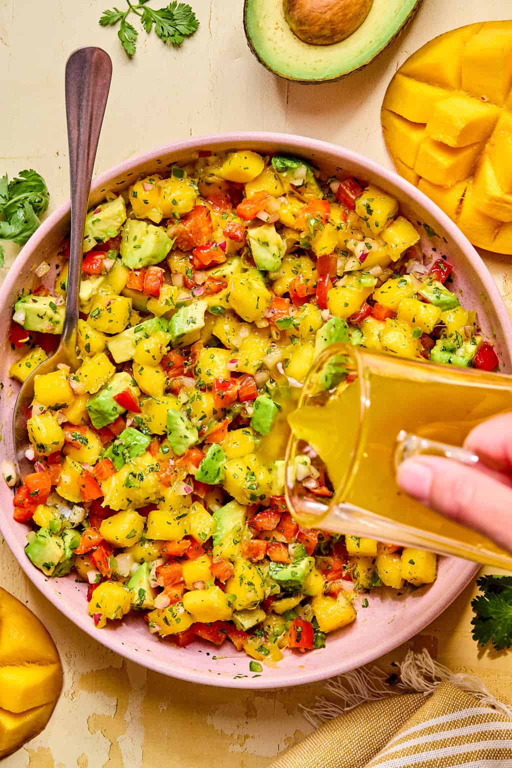 A bowl of vibrant mango avocado salad with diced red bell pepper and herbs, while a hand pours dressing from a glass. Half an avocado and sliced mango rest nearby on a yellow surface.